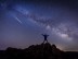 Long exposure image including: bright shooting star (pressed, meteor) next to the beautiful Milky Way Galaxy and countless stars on a clear summer starry night as seen from asia's Northern hemisphere. Silhouettes of man and hill as foreground to frame the shot. Light pollution evident on the horizon from nearby towns and human settlements. Shot on Nikkor D4s,lens by 14-24.